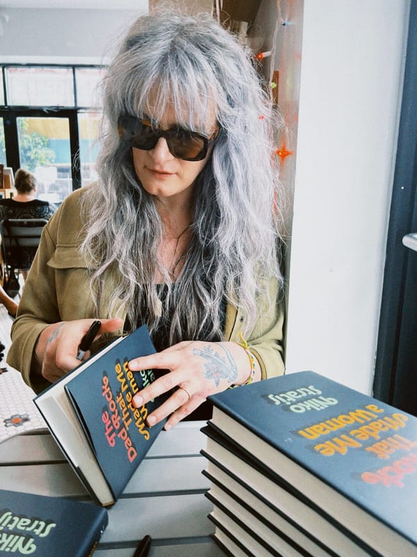 a woman with grey hair and a green jacket signs copies of a book called The Dad Rock That Made Me a Woman